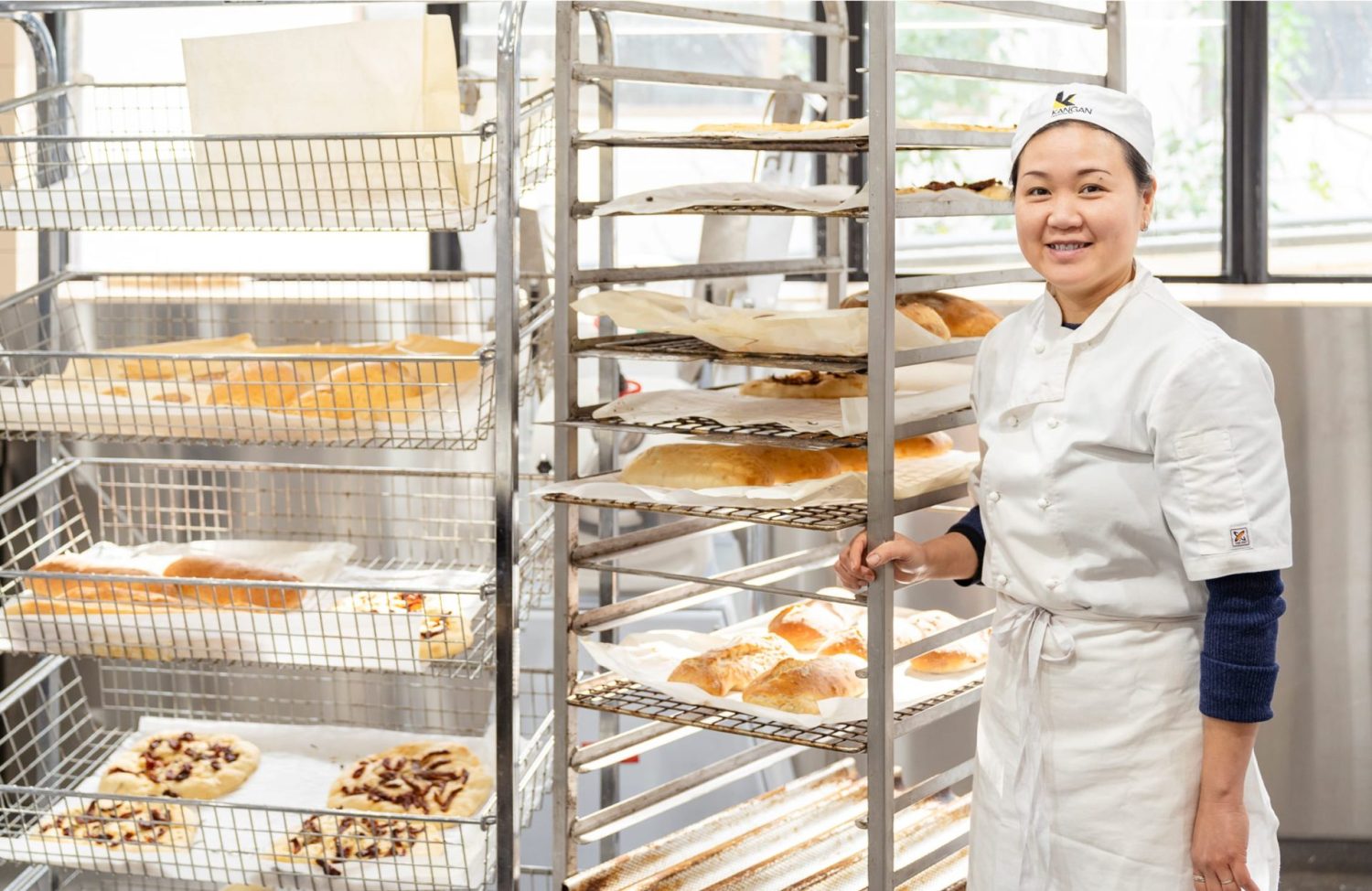 A woman in a baker's uniform stands smiling next to a rack filled with freshly baked bread and pastries.