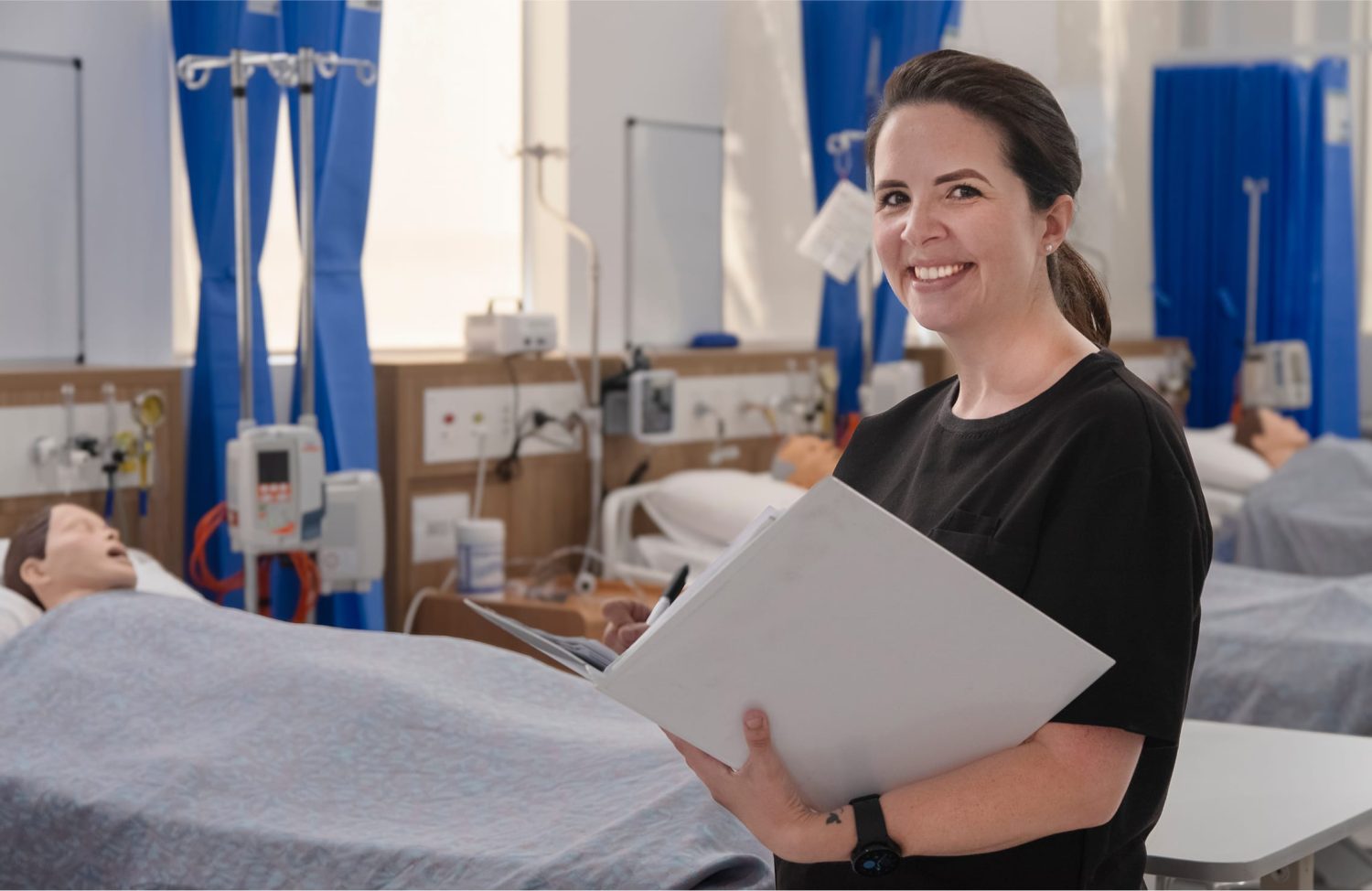 A female student in a black shirt holds a folder and smiles in a simulated hospital ward with mannequins in beds behind her.