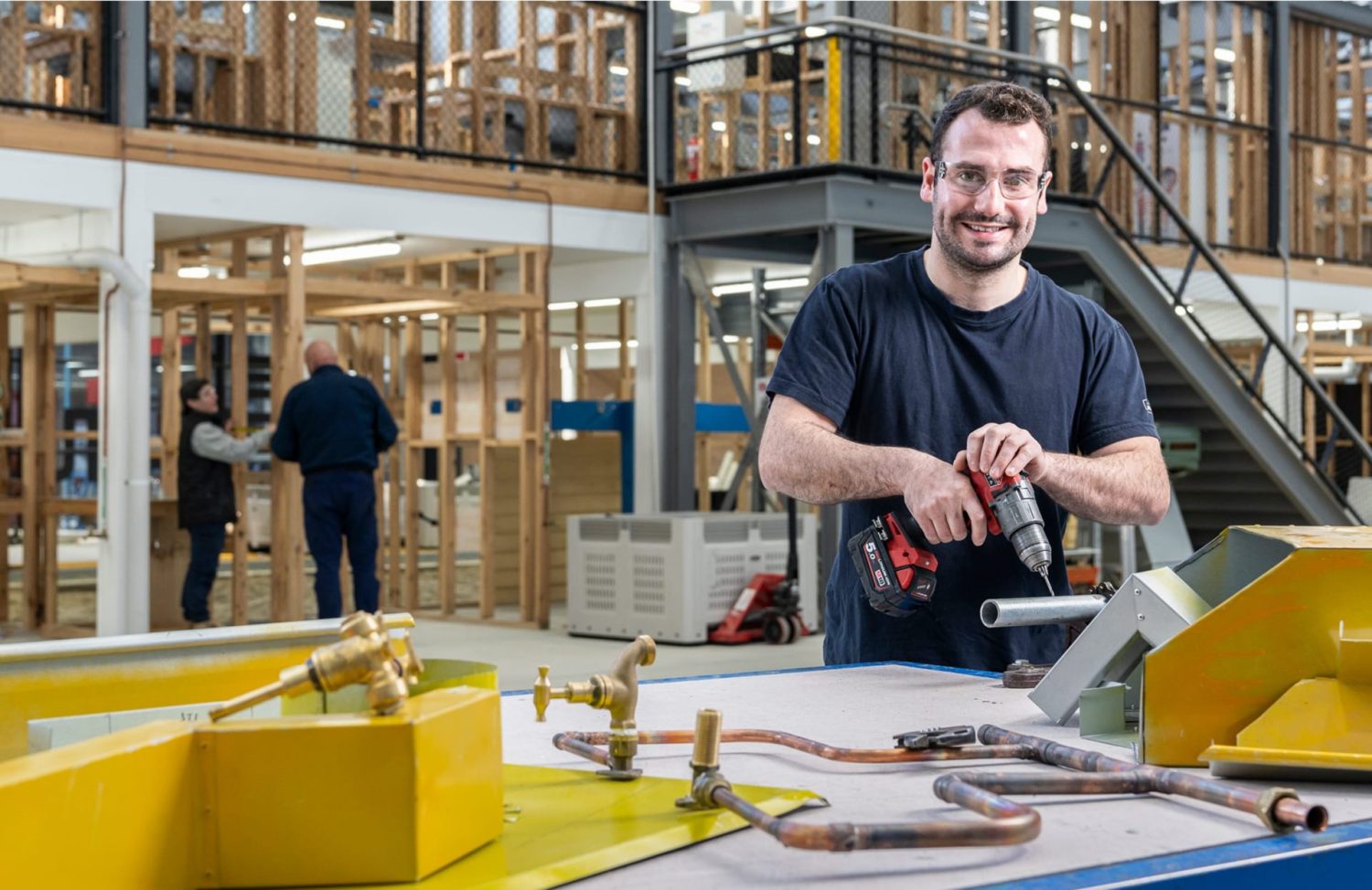 A male student in a workshop holds a drill while smiling at the camera, with a bench of pipes and tools in the foreground.