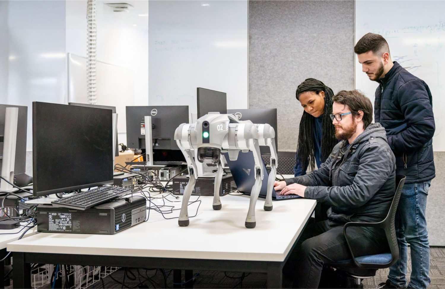 A group of three students, two men and one woman, gather around a table with a laptop and a grey four-legged robot.