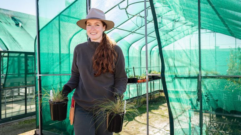 A horticulture student, wearing a hat, dark clothes, and gloves, smiles while holding two small potted plants inside a large green shade cloth greenhouse.
