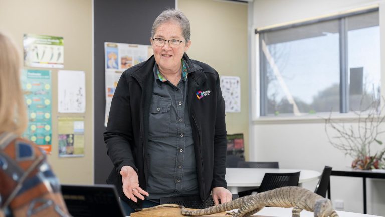 A teacher, in a black jacket and glasses, gestures while speaking to a student in a classroom
