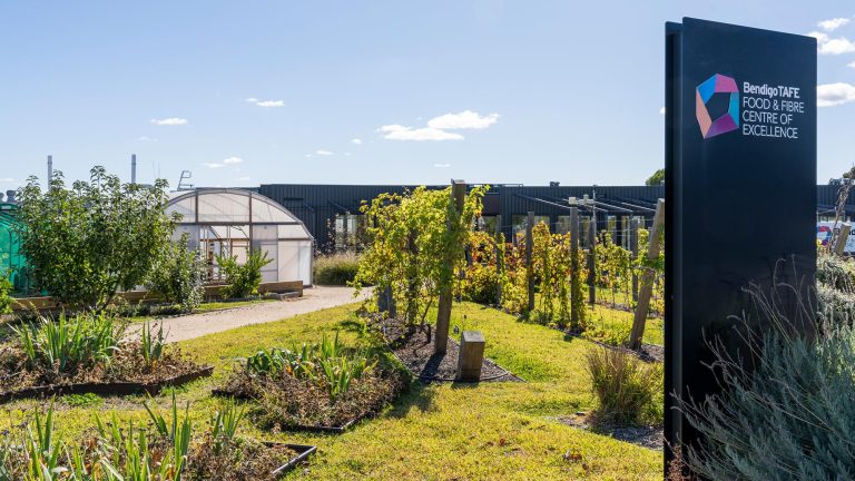 Exterior view of the Bendigo TAFE Food & Fibre Centre of Excellence, showing a black signage monolith in the foreground, a landscaped garden with a small greenhouse, and the main campus building in the background.