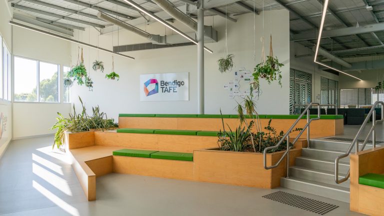 Bright, modern interior entrance area of the Bendigo TAFE Food & Fibre Centre of Excellence, featuring tiered wooden seating with green cushions, integrated planters with lush plants, and a Bendigo TAFE banner on the wall.