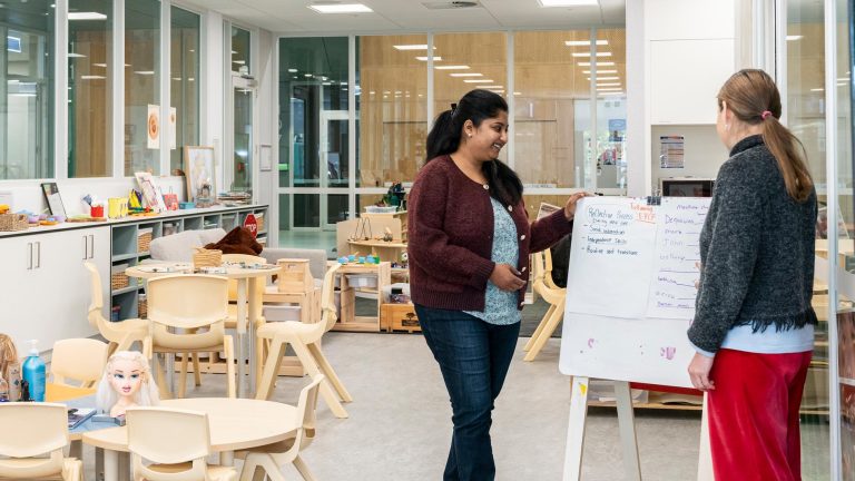 An interior of an early childhood education classroom or play space. A female teacher, dressed in a maroon cardigan, stands next to a whiteboard explaining a concept to a student. The room has light wood shelving, small tables and chairs suitable for children, and various toys and learning materials.