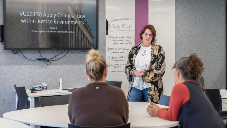 A classroom setting where a female teacher stands in front of a whiteboard and a large screen addressing two students seated at a round table