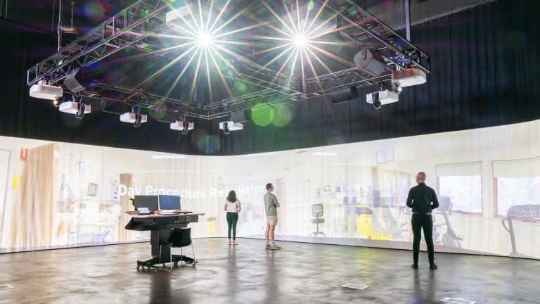 A wide shot of an immersive learning gallery or simulation room. The walls are a curved projection screen displaying a hospital ward or procedure room. Four people stand in the room, two of them facing a central monitoring station with computers, under an array of overhead lights mounted on a truss structure.