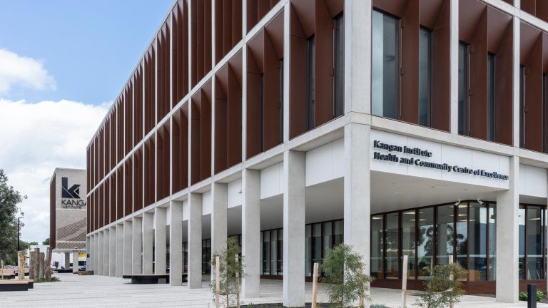 A modern, multi-story building exterior featuring a facade of brown vertical fins. The entrance, supported by large white concrete columns