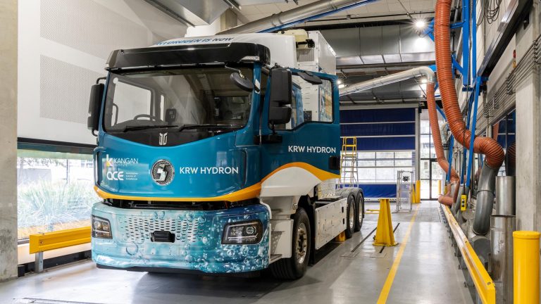 A large, blue and white truck, branded "parked inside a high-tech workshop bay with visible exhaust extraction hoses overhead.