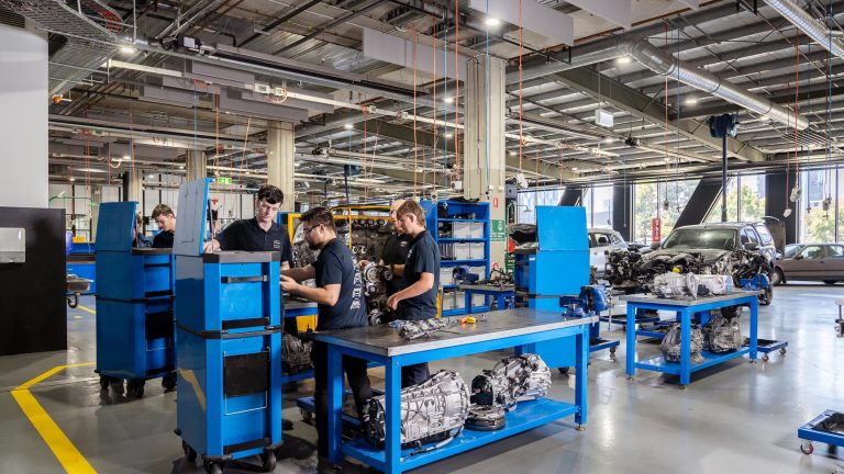 A group of light vehicle mechanical students are gathered around workbenches with engine components in a large, well-equipped workshop featuring blue tool cabinets and yellow-lined floors.