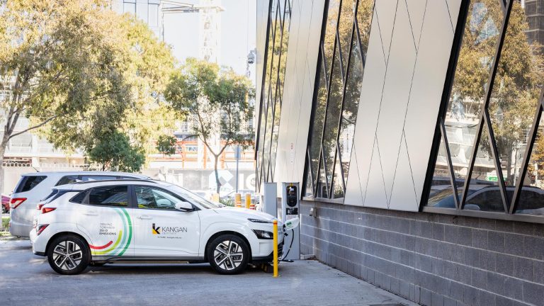 A white vehicle, branded with the Kangan Institute logo is parked and charging at an electric vehicle charging station outside a modern building with an angled facade
