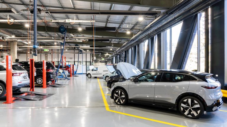 Interior view of a brightly lit automotive workshop at Kangan Institute Docklands campus, showing several vehicles on lifts and a new gray electric vehicle (EV) parked in the foreground with its hood open, showcasing the modern facility's high windows.