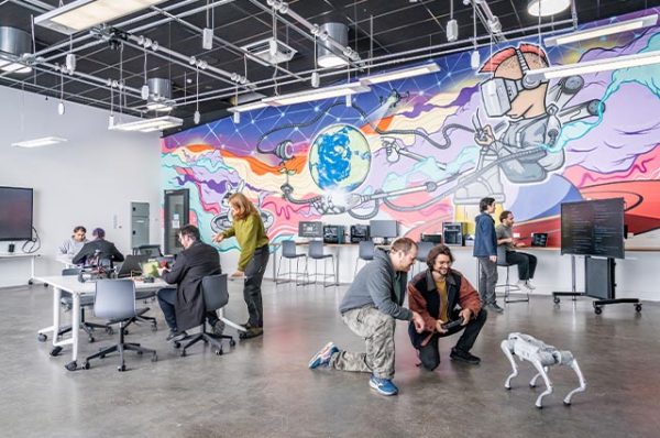 Interior of a brightly lit digital workshop space at Kangan Institute Cremorne, featuring a colorful mural on the wall, and students or instructors working on laptops and interacting with a small robotic dog.
