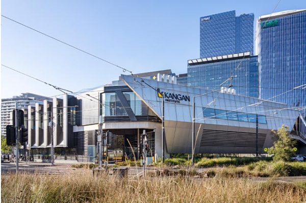 Exterior of the modern Kangan Institute Docklands Campus building, featuring a distinct, angular metal facade, set against a backdrop of tall corporate skyscrapers and tram lines in the foreground.