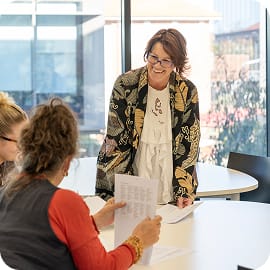 A candid indoor shot of a woman with short dark hair and glasses standing and smiling brightly over a round white table, interacting with two people seated across from her