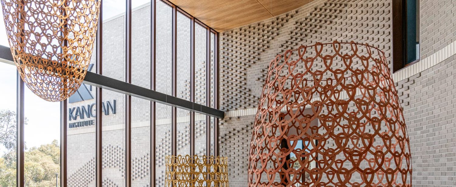 An architectural photograph of a modern interior space, featuring a light grey textured brick wall with a distinctive patterned recess. In the foreground, there are large, decorative, cone-shaped light fixtures or sculptures made of woven metal circles in rusty orange/brown and yellow/gold tones.