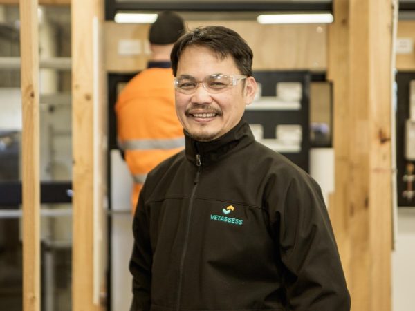 A medium shot of a man named Michael smiling at the camera. He is wearing a black zippered jacket with the "VETASSESS" logo on the chest and clear safety glasses.