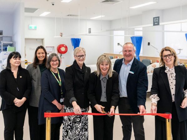 A group of seven people, standing in a brightly lit hospital or medical training room, performing a ribbon-cutting ceremony.