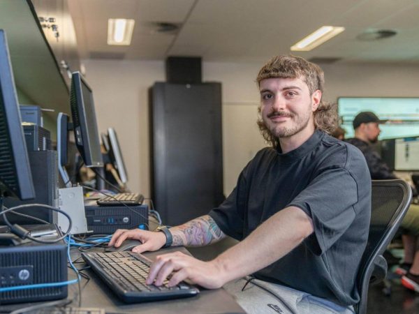 A young man with a mustache and a prominent mullet hairstyle, sitting at a computer desk in an office environment. He is looking at the camera and smiling faintly.