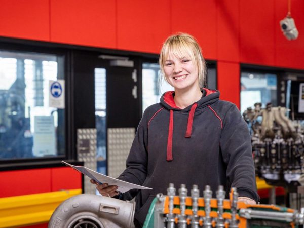 A young woman smiling brightly at the camera. She has blonde hair with bangs and is wearing a dark grey/black hooded sweatshirt with red trim. She is holding a notebook and pen. She is in a workshop or industrial setting, with a large piece of machinery in the foreground and a red wall in the background.