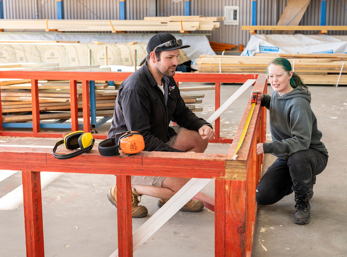 Two people in a workshop or warehouse setting, crouched down, measuring a wooden frame with a yellow tape measure.