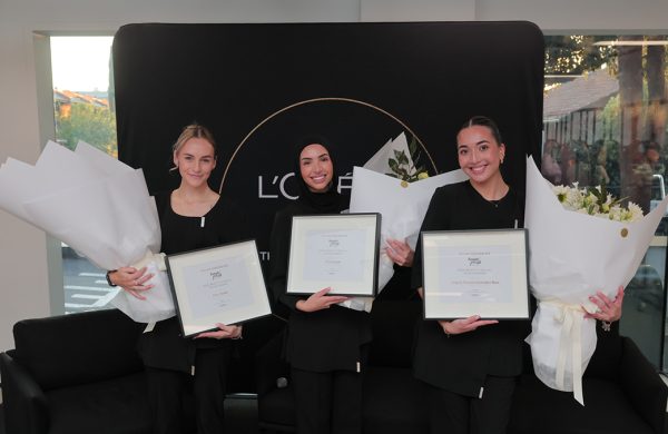 Three beauty scholars holding certificates and bouquets of flowers, smiling in front of a Loreal sign